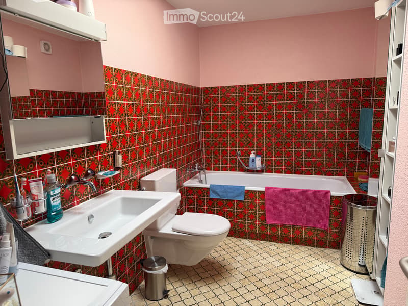 Bathroom with pink walls, red and black patterned tiles, a white sink, toilet, bathtub, and various toiletries.