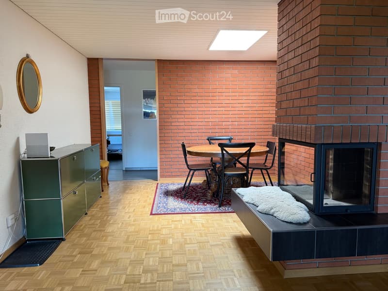 Modern living room with a fireplace, wooden floor, round dining table with chairs, a cabinet with a laptop, and a decorative mirror on the wall.