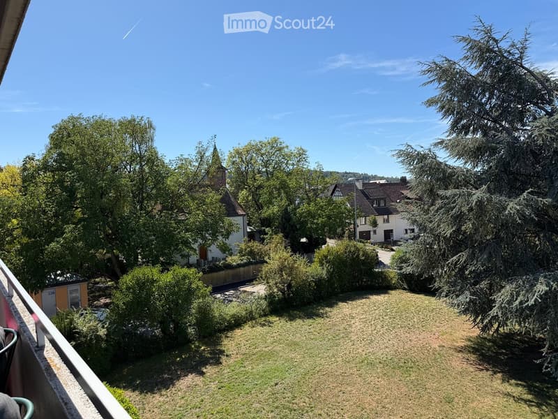 View from a balcony showing a well-maintained garden with grass, bushes, and trees, with neighboring houses and a church visible in the background under a clear blue sky.