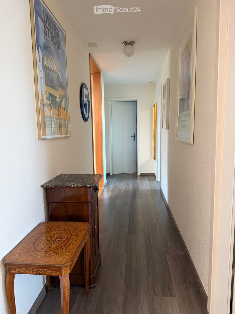 A narrow hallway with wooden flooring, white walls, and several framed artworks. A small wooden table and cabinet are placed on the left side.