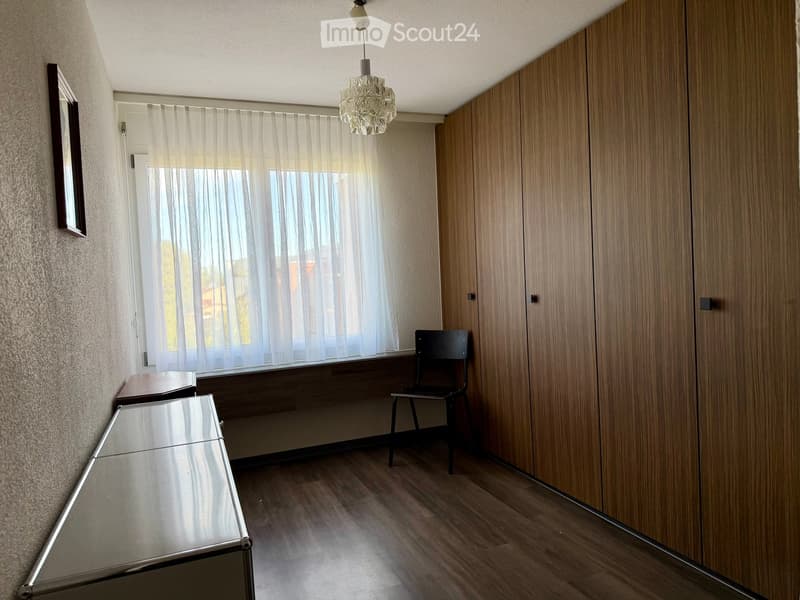 A narrow hallway with wooden flooring, a large window with sheer curtains, a modern pendant light, a small table, a chair, and a large mirrored cabinet.