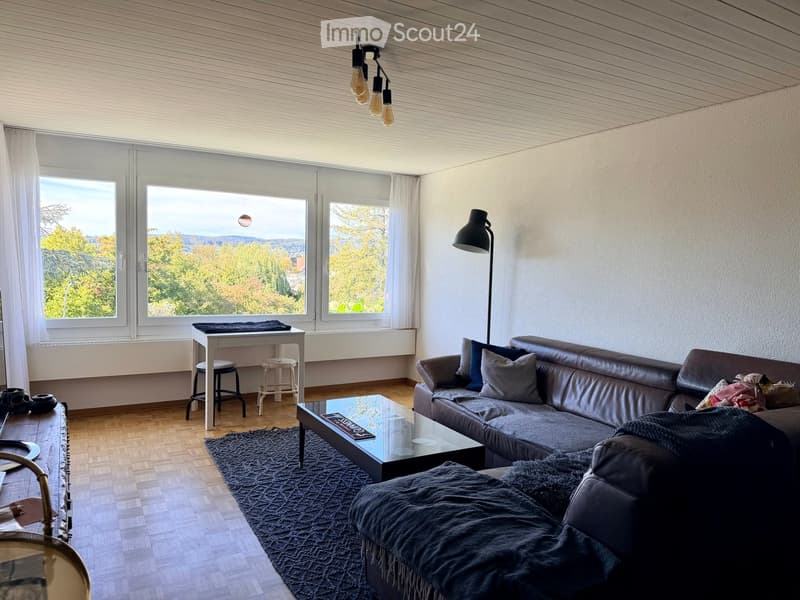 Modern living room with large windows offering a scenic view, featuring a dark leather sofa, glass coffee table, small table with stools, and a floor lamp.