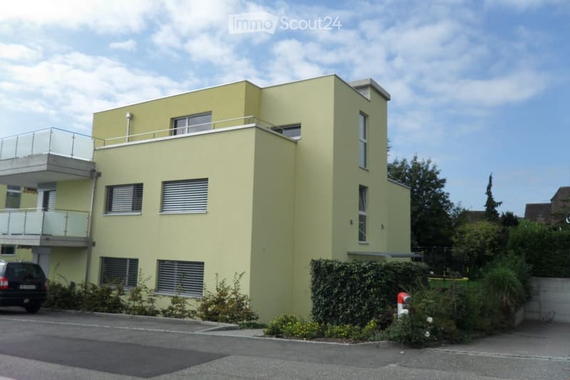 Modern two-story building with light yellow facade, large windows, and balconies. Features include a raised ground floor, parking area, and landscaped surroundings.