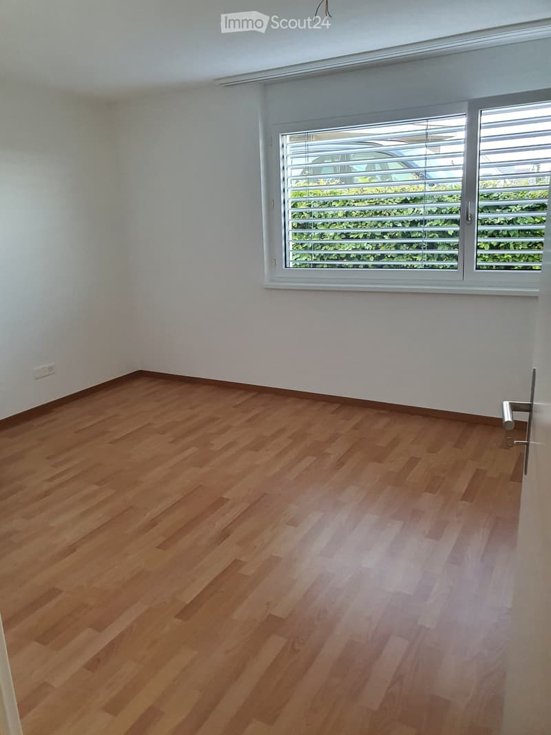 Empty room with wooden flooring and white walls, featuring a large window with blinds and a door handle visible.