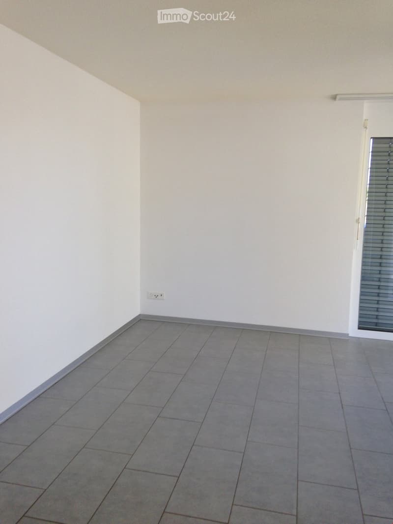 Empty room with tiled floor and white walls, featuring a power outlet and a partially visible door with blinds.