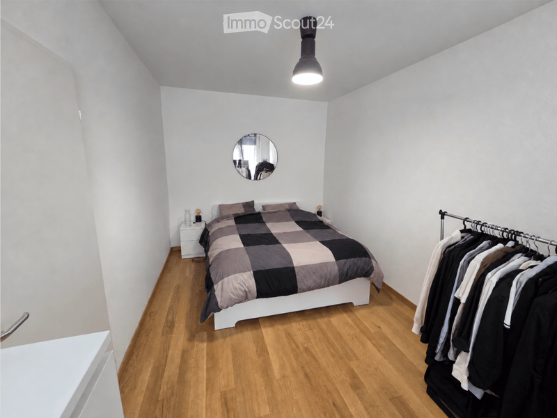 Modern bedroom with wooden flooring, white walls, a bed with a checkered duvet, a nightstand, a round mirror, and a clothing rack with hanging garments.