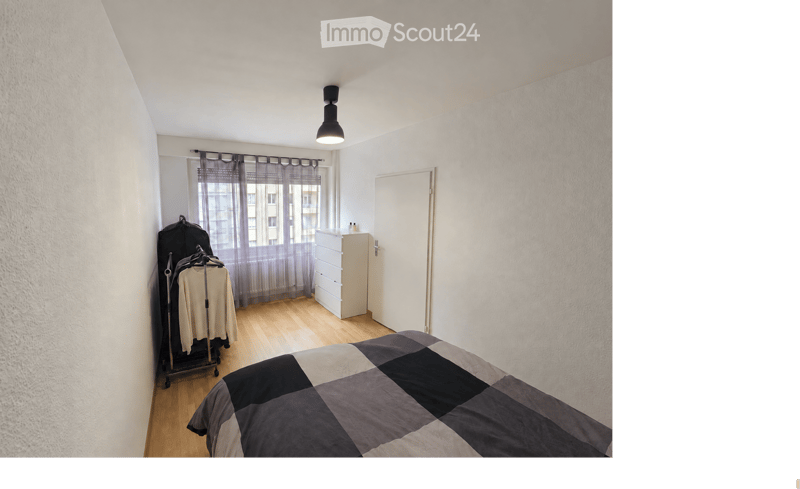 A modern bedroom with a wooden floor, a bed with a grey and black checkered duvet, a white wardrobe, a clothing rack with hanging clothes, and a window with grey curtains.