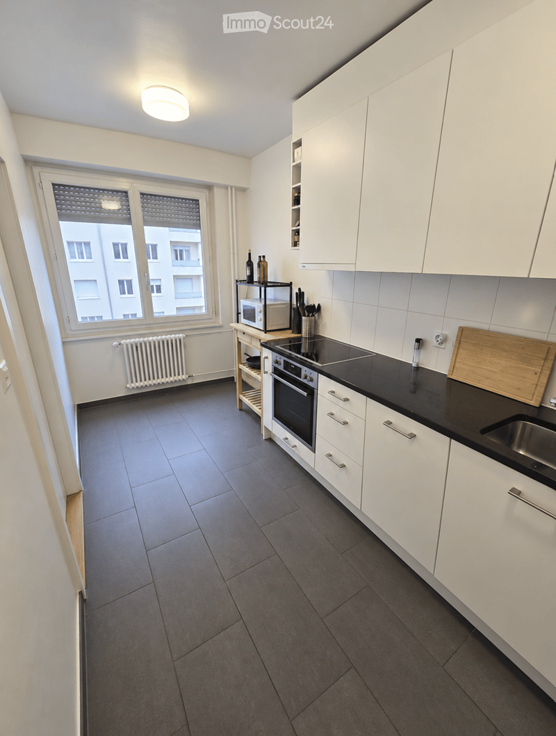Modern kitchen with white cabinets, black countertops, and stainless steel appliances. Features an oven, microwave, sink, and a small wooden shelf with bottles and utensils.
