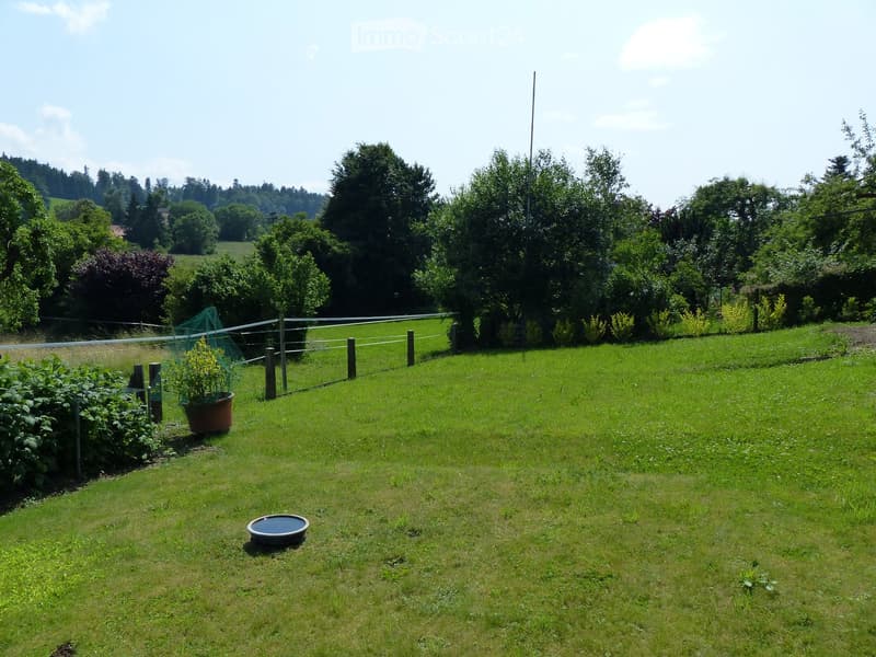 Well-maintained lawn with a fence, a few potted plants, and a small water dish, surrounded by trees and greenery.
