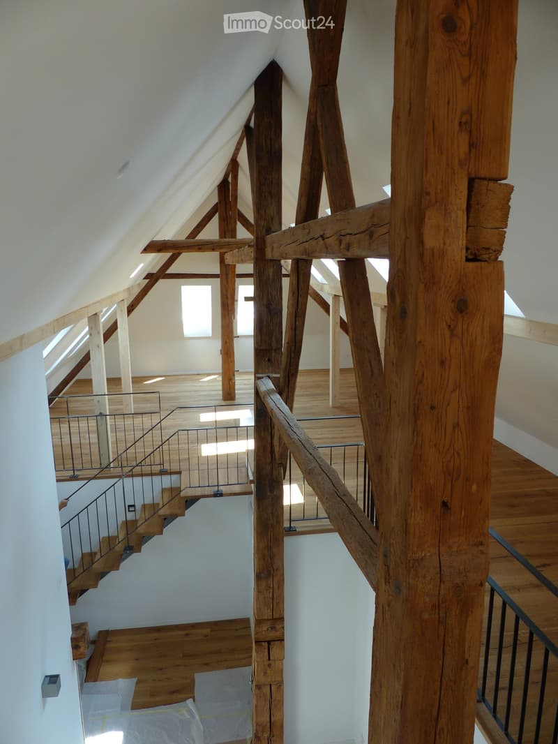 Spacious, open hallway with wooden beams and a staircase leading to an upper level, featuring wooden flooring and large windows allowing natural light to fill the space.