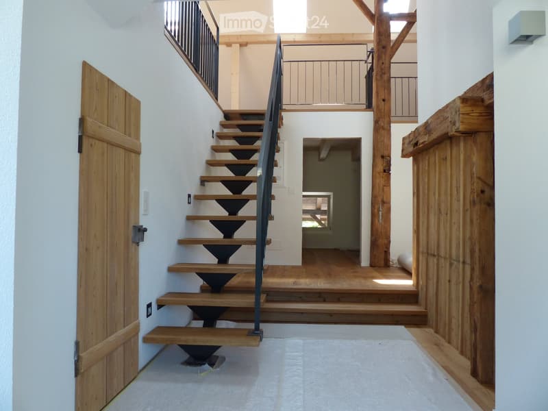 Modern hallway with wooden staircase, white walls, and wooden doors. The staircase features wooden steps and a metal railing, leading to an upper level with visible wooden beams and a railing.