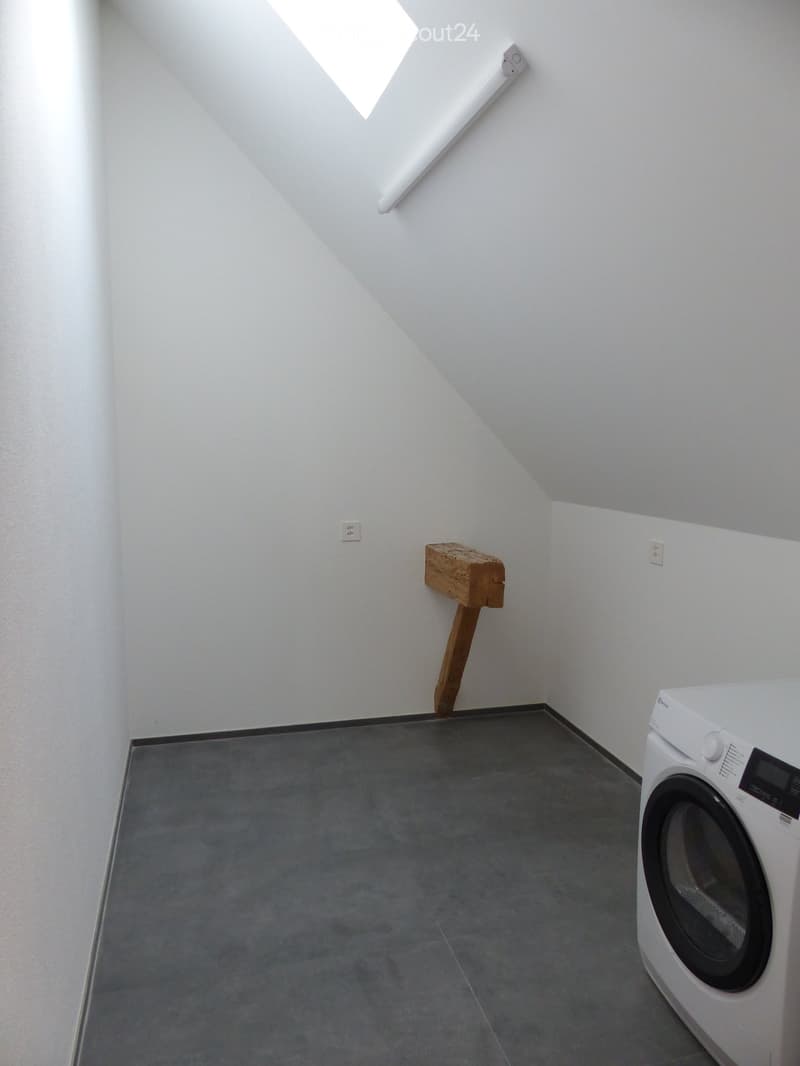 Empty room with white walls, grey floor, and a white washing machine in the corner. A small wooden shelf is mounted on the wall.