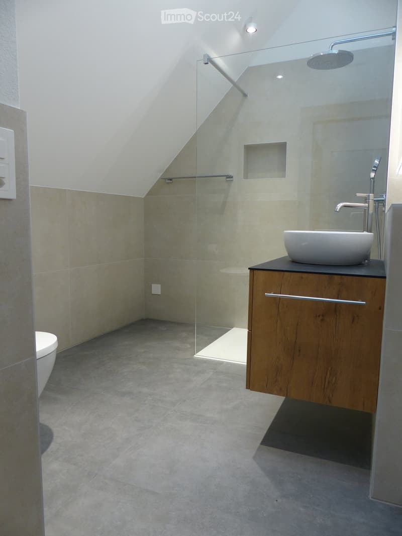 Modern bathroom with a sleek sink, faucet, toilet, and a glass-enclosed shower area. The space features light-colored tiles and a wooden cabinet.