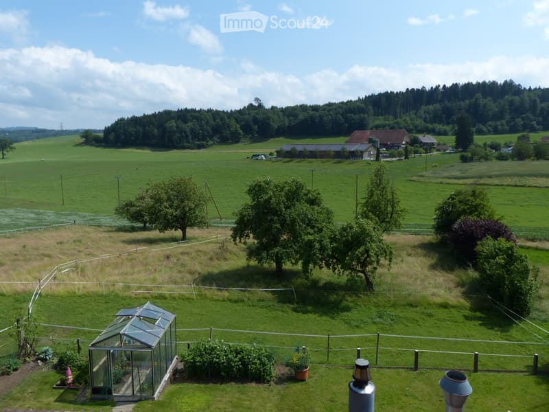 A peaceful garden area with a greenhouse, fenced perimeter, trees, and a scenic view of a rural landscape with fields and houses in the distance.