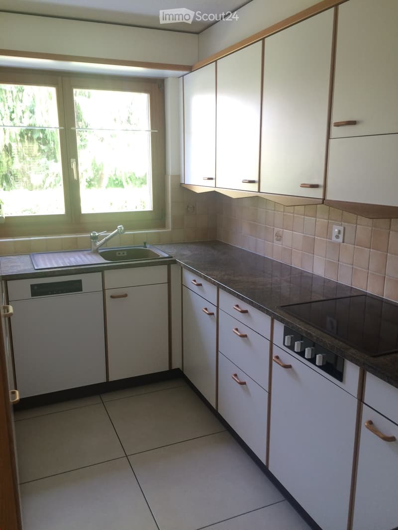 Modern kitchen with white cabinets, beige tiles, and a window overlooking greenery. Features a sink, faucet, dishwasher, and stove.