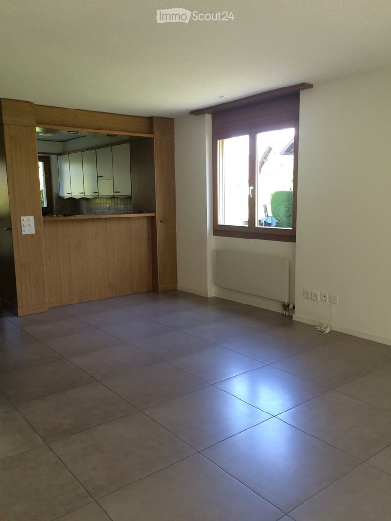 Empty room with tiled floor, light-colored walls, a large window with wooden frame, radiator, and a wooden kitchen counter visible in the background.