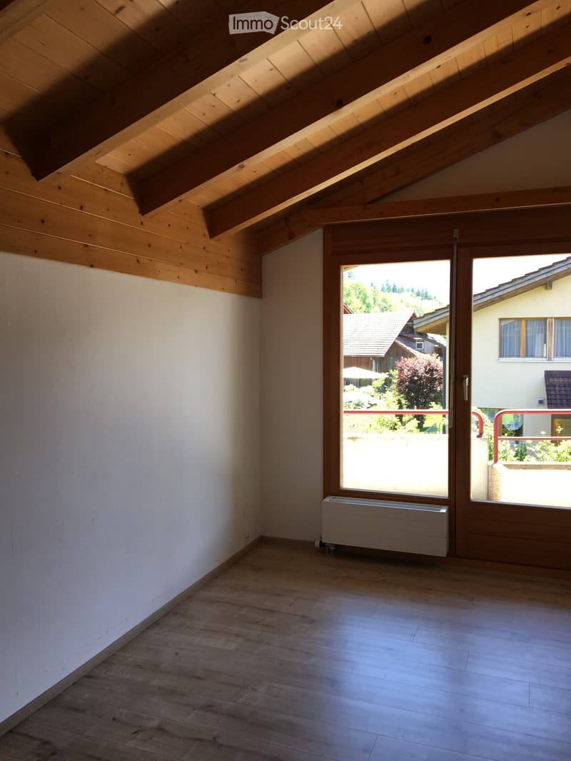 Empty attic room with wooden slatted ceiling, light wooden flooring, white walls, and a large window offering a view of the surrounding landscape.