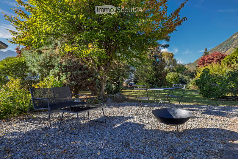 Garden area with gravel ground, featuring a black bench, chairs, a small table, and a fire pit, surrounded by trees and greenery with a mountain view in the background.