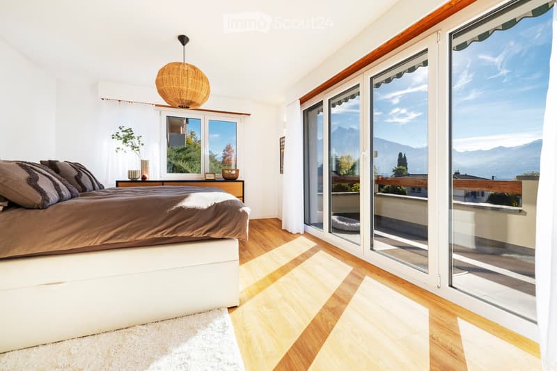 Bright bedroom with wooden flooring, large windows offering a scenic mountain view, a neatly made bed with pillows and blankets, a nightstand with a lamp, and a small plant on the windowsill.