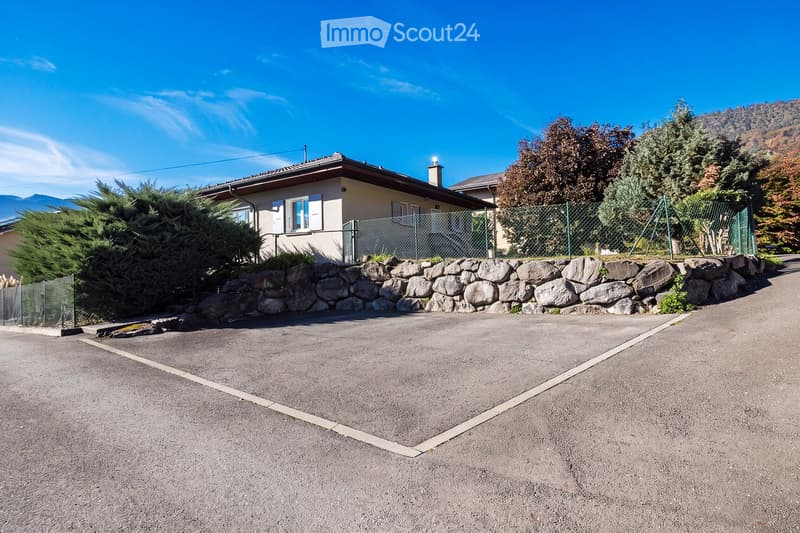 A two-story house with a stone wall and a fenced yard, surrounded by trees and mountains under a clear blue sky.