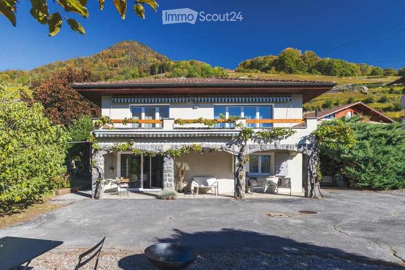 Two-story house with a stone lower level and a wooden upper level, surrounded by greenery and a scenic mountain view.