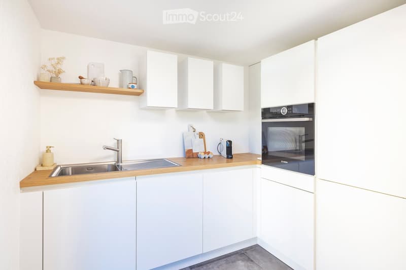 Modern kitchen with white cabinets, wooden countertops, stainless steel sink, and built-in oven. Shelves hold decorative items and kitchenware.