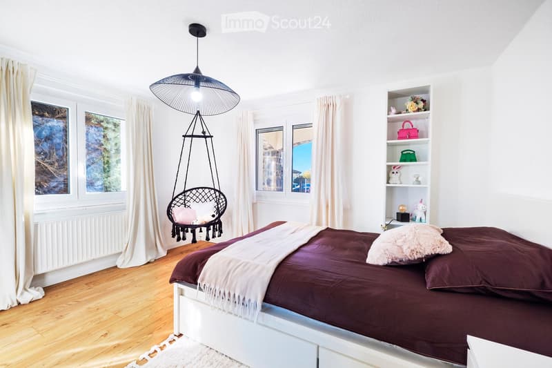 Cozy bedroom with wooden flooring, large windows, a white bed with dark bedding, a hanging wicker chair, and shelves with decorative items.
