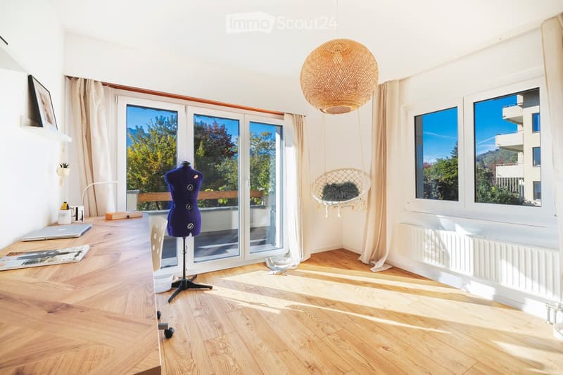 Bright, airy room with wooden flooring, large windows, and a balcony view. Features include a hanging wicker chair, a mannequin, a white desk chair, and a laptop on a table.