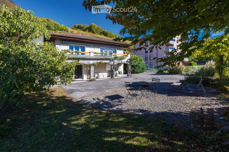 A well-maintained garden area with a gravel patio, outdoor furniture, and a stone house with a balcony and large windows.