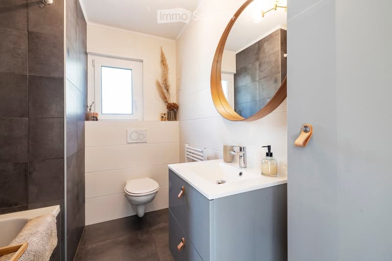 Modern bathroom with a white sink, chrome faucet, round wooden-framed mirror, toilet, bathtub, and decorative items on the windowsill.