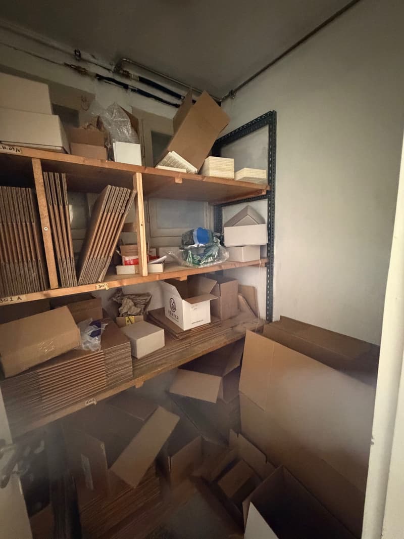A storage room filled with various cardboard boxes, some stacked on shelves and others on the floor. The room has white walls and a metal rack on the right side.
