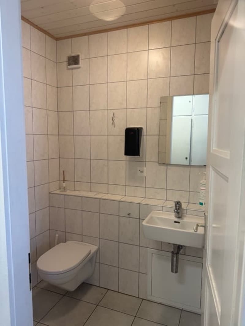 Bathroom with white tiles, toilet, sink, mirror, and soap dispenser.