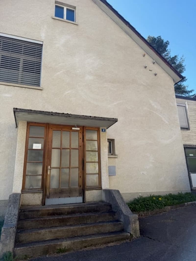 A beige building with a wooden door and glass windows. The building has a slanted roof and a small balcony on the second floor. The building has a number 7 on the wall above the door.