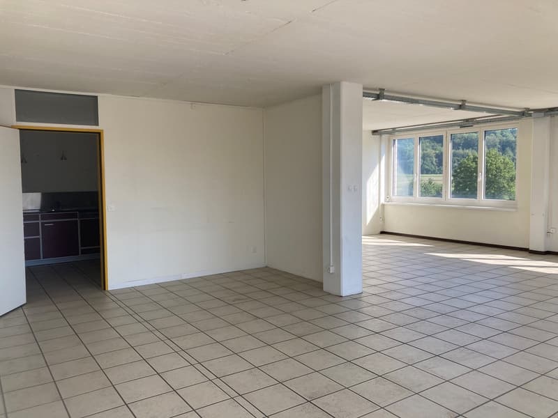 Empty room with tiled floor, white walls, open door leading to a small kitchenette with cabinets and windows.