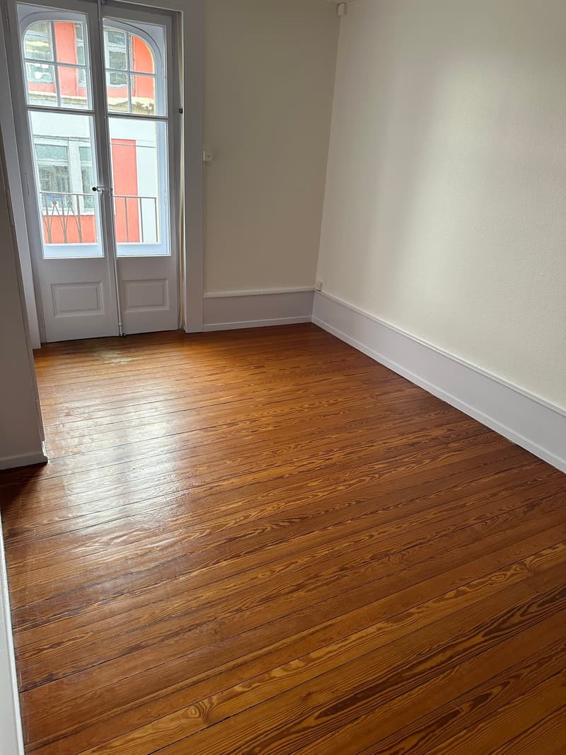 Empty room with hardwood floors, white walls, large doors, window overlooking balcony.