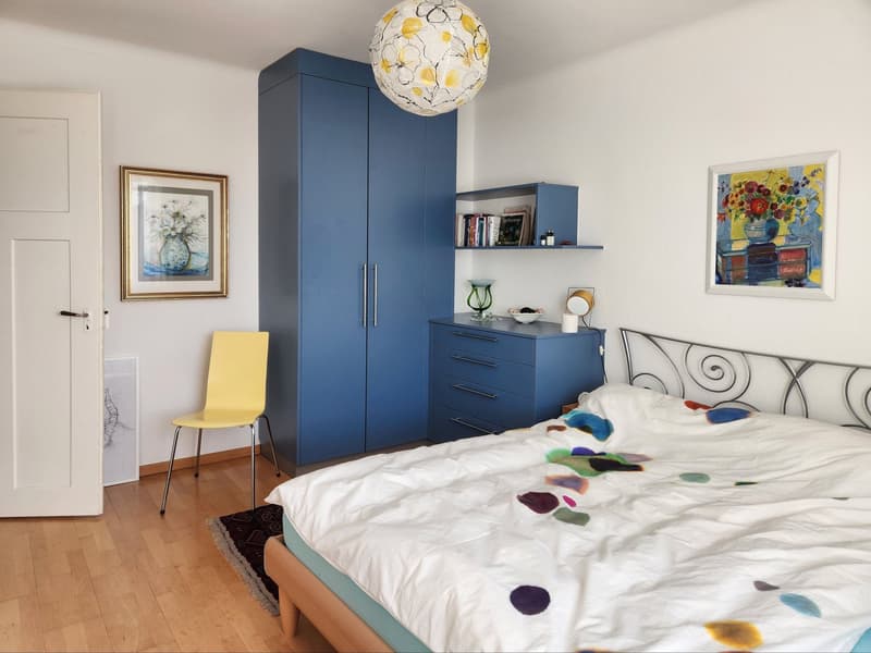 Bedroom with bed, blue wardrobe, yellow chair, wooden floor, white walls, paintings, shelf, and nightstand
