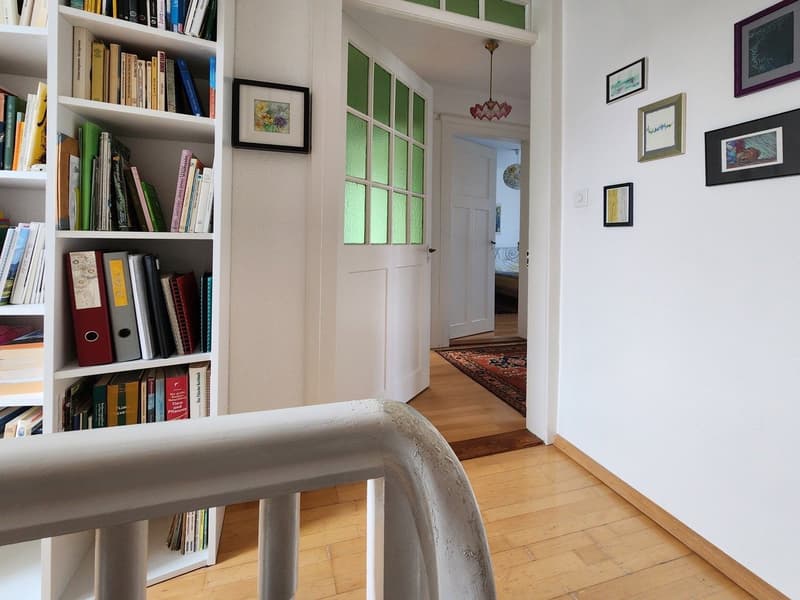 A hallway with a wooden floor, white walls, a white bookshelf filled with books, a white door with green glass panels, a rug, and framed pictures on the wall.