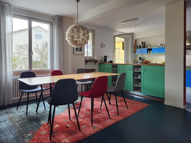 Dining room with a long wooden table, colorful chairs, a red rug, and a chandelier. The room has large windows with white curtains, a radiator, and a green kitchen with cabinets and shelves.