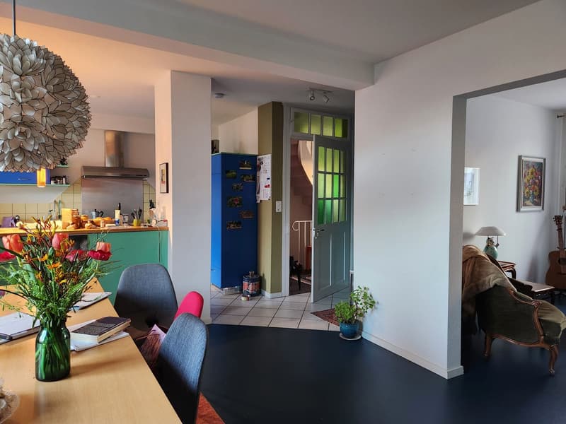 Dining room with a table, chairs, and a kitchen in the background. A lamp, a plant, and a guitar are also visible.