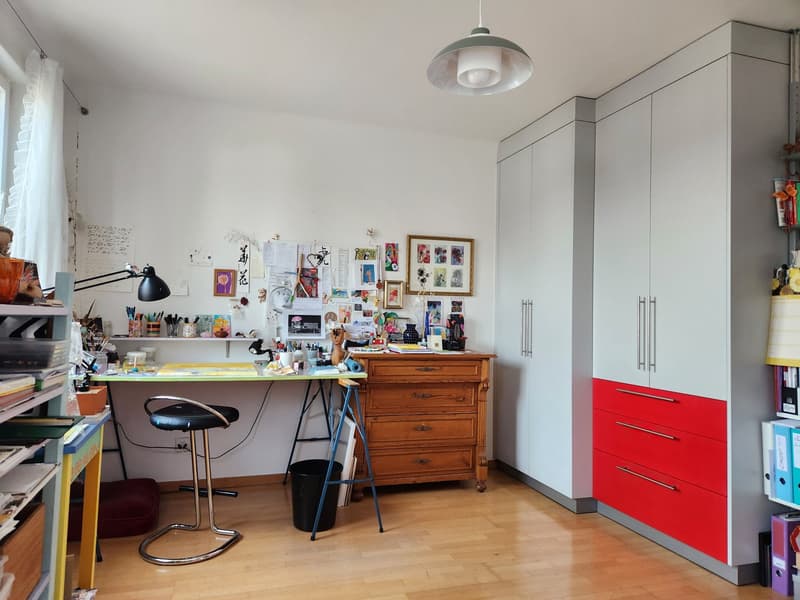 A room with a desk, a stool, a cabinet, a shelf, a lamp, a trash can, a wall with pictures, and a window with a curtain.