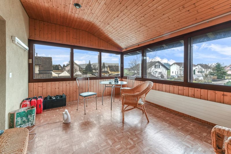A sun room with wooden walls and floor, a small round table with two chairs, and large windows overlooking a neighborhood