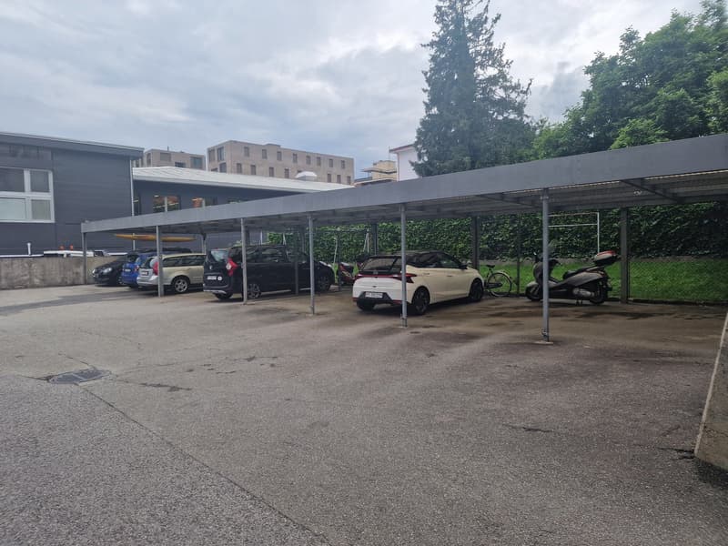 covered parking lot with several cars parked, metal poles, metal roof, green grass, buildings in the background