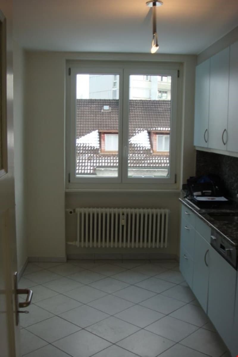 A kitchen with a large window, a radiator, white tiled floor, cabinets, countertop, and an open door.