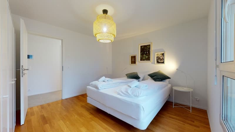 Bedroom with white bed, wooden floor, white walls, yellow lamp, white nightstand, white towels, white sheets, green pillows, framed pictures on wall, open door.