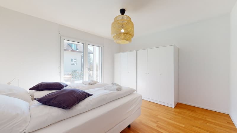 Minimalist design, white walls, wooden floors, double bed with purple pillows, white cabinet, glass door with view of outside.