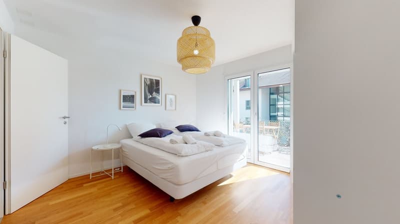 a double bed with pillows and white sheets, a small white side table with a lamp, a wall with a picture frame, a wooden floor, a white door, a glass door that leads to a balcony