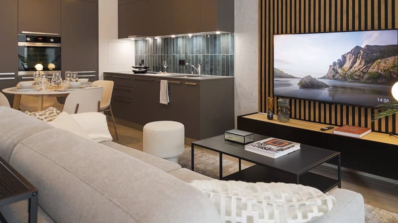 The image shows an open-concept living room and kitchen area. The kitchen features dark cabinets, a tile backsplash, and stainless steel appliances. The living room has a large sofa, a coffee table, and a TV mounted on the wall. The room also has a large