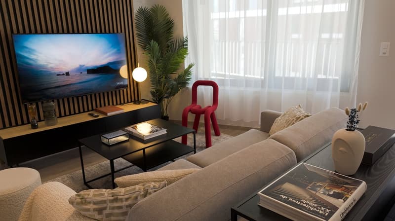 Cozy living room with large windows, a beige sofa, a black coffee table, and a framed artwork depicting a scenic beach sunset. The room also features a red sculptural chair and potted plants, creating a modern and relaxing atmosphere.