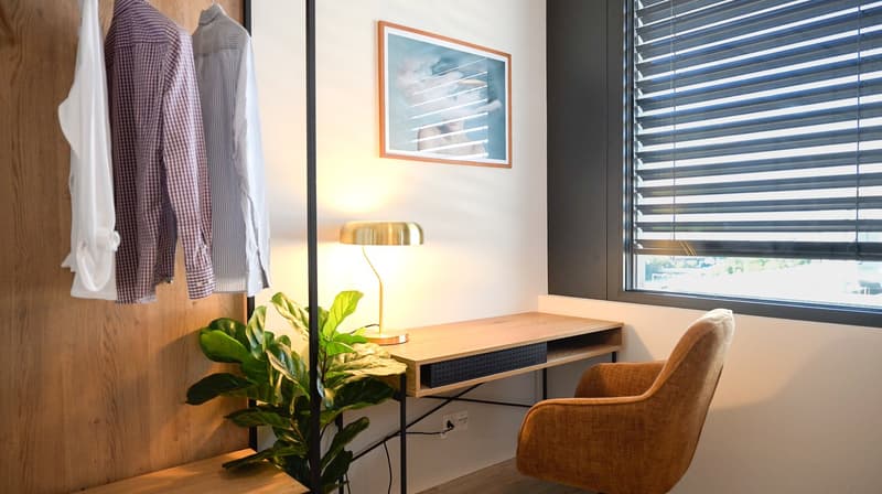 Wooden desk with storage, orange chair, potted plant, framed artwork, and blinds on the window