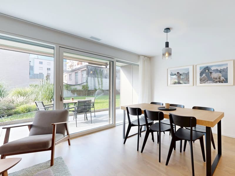 Modern dining room with a large wooden table, black chairs, and sliding glass doors leading to a balcony. Picture frames on the wall and a lamp hanging from the ceiling. View of an outdoor garden with a table and chairs. Light-colored flooring and a carpe